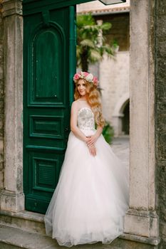 A bride with a wreath of roses stands at the green door in the cozy courtyard of the old town of Kotor . High quality photo