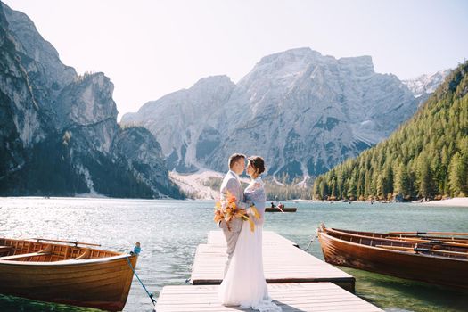 The bride and groom walk on a wooden boat dock at Lago di Braies in Italy. Wedding in Europe, at Braies lake. Newlyweds walk, kiss, cuddle against the backdrop of rocky mountains.