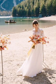 Beautiful bride in a white dress with sleeves and lace, with a yellow autumn bouquet against backdrop of arch for ceremony, at Lago di Braies in Italy. Destination wedding in Europe, at Braies lake.