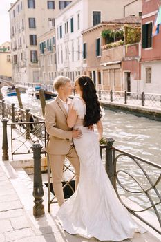 Newlyweds stand hugging on the banks of the Venice Canal. The groom hugs the bride by the waist. White wedding dress with a small beautiful train and a sandy color men's suit.