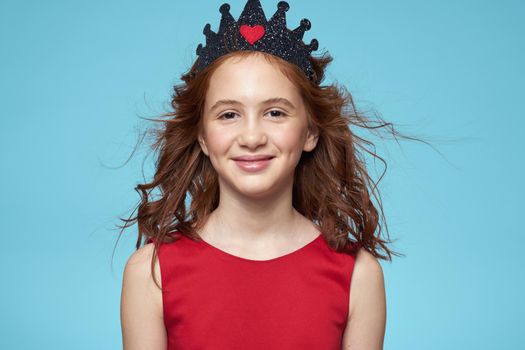 little girl with curly hair with a crown on her head red dress Smile blue background. High quality photo