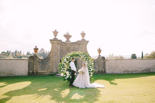 Wedding couple near round wedding arch decorated with white flowers and greenery in front of an ancient Italian architecture. Wedding at an old winery villa in Tuscany, Italy