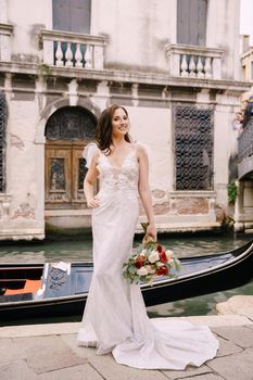 A bride in a white dress, with a train, with a bouquet of white and red roses in her hands, stands on the pier near the moored gondola in a narrow Venetian canal.