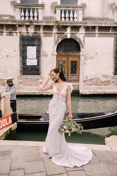 A bride in a white dress, with a train, with a bouquet of white and red roses in her hands, stands on the pier near the moored gondola in a narrow Venetian canal.