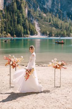 Beautiful bride in a white dress with sleeves and lace, with a yellow autumn bouquet against backdrop of arch for ceremony, at Lago di Braies in Italy. Destination wedding in Europe, at Braies lake.