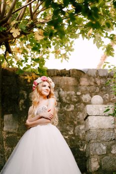 A bride in a delicate wreath stands by a brick wall under a fig tree, her arms folded on her chest . High quality photo