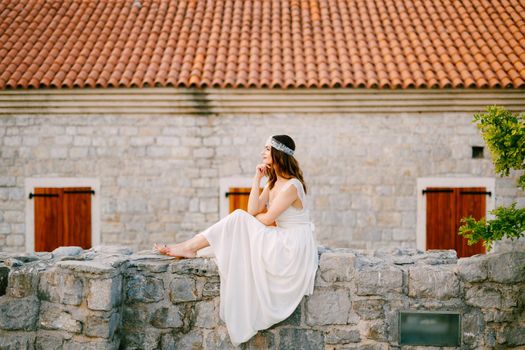 Barefoot bride sits on the stone wall of the old town of Budva . High quality photo