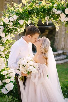 Wedding at an old winery villa in Tuscany, Italy. Round wedding arch decorated with white flowers and greenery in front of an ancient Italian architecture. The wedding couple is kissing.