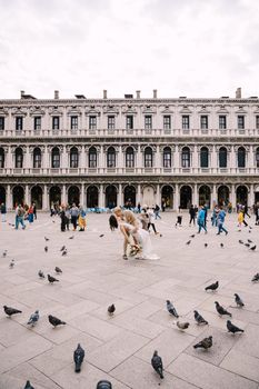 Wedding in Venice, Italy. The bride and groom kiss among the many pigeons in Piazza San Marco, against backdrop of the National Archaeological Museum Venice, surrounded by a crowd of tourists.