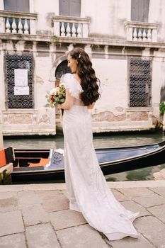 A bride in a white dress, with a train, with a bouquet of white and red roses in her hands, stands on the pier near the moored gondola in a narrow Venetian canal.