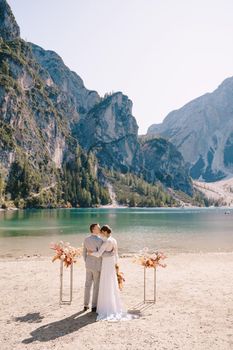 The bride and groom stand still for the ceremony, with an arch of autumn floral columns, against the backdrop of Lago di Braies in Italy. Destination wedding in Europe, at Braies lake.