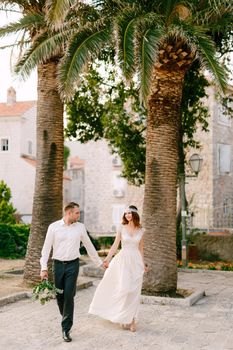 The bride and groom walk hand in hand through the old town of Budva, next to beautiful tall palm trees . High quality photo