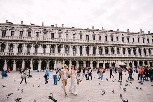 Wedding in Venice, Italy. The bride and groom are running through a flock of flying pigeons in Piazza San Marco, amid the National Archaeological Museum Venice, surrounded by a crowd of tourists.