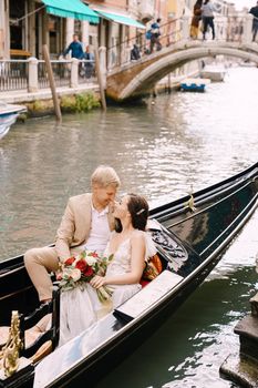 The bride and groom ride in a classic wooden gondola along a narrow Venetian canal. A close-up of cuddling newlyweds.