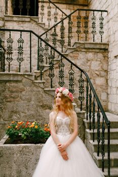 A bride with a wreath of white and pink roses stands near a staircase with a wrought railing in the old town of Kotor . High quality photo