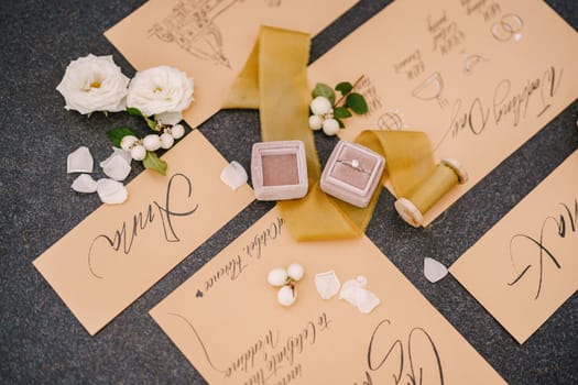 Wedding rings in a box, invitation cards, silk ribbon, white roses on black background. Overhead top view
