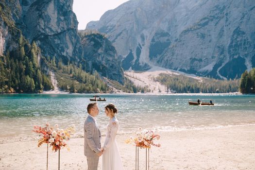 The groom dons a bride's ring, in place for the ceremony, with an arch of autumn floral columns, against the backdrop of Lago di Braies in Italy. Destination wedding in Europe, at Braies lake.