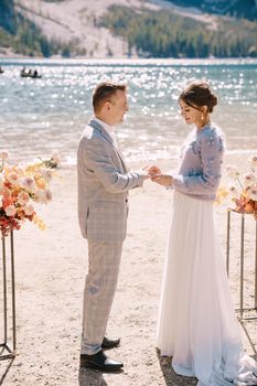The groom dons a bride's ring, in place for the ceremony, with an arch of autumn floral columns, against the backdrop of Lago di Braies in Italy. Destination wedding in Europe, at Braies lake.