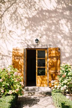 Old wooden door open and the facade of the villa in Italy under the shade of trees, hydrangea flower pots illuminated by the sun.