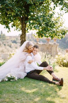 The wedding couple sits on the grass in the garden under a tree, the bride hugs the groom. Wedding in Florence, Italy, in an old villa-winery