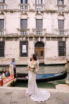 A bride in a white dress, with a train, with a bouquet of white and red roses in her hands, stands on the pier near the moored gondola in a narrow Venetian canal.
