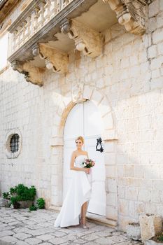 A bride in an elegant dress with a bouquet in her hands stands at the white doors of a beautiful old building in Perast . High quality photo