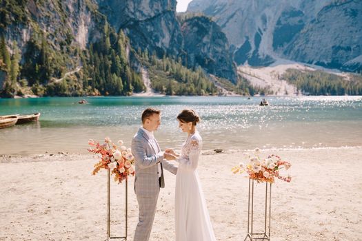 The bride dons a ring to the groom at the venue for the ceremony, with an arch of autumn floral columns, against the backdrop of Lago di Braies in Italy. Destination wedding in Europe, at Braies lake.
