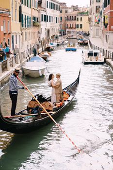 The gondolier rides the bride and groom in a classic wooden gondola along a narrow Venetian canal. The newlyweds stand in the boat, the view from behind.