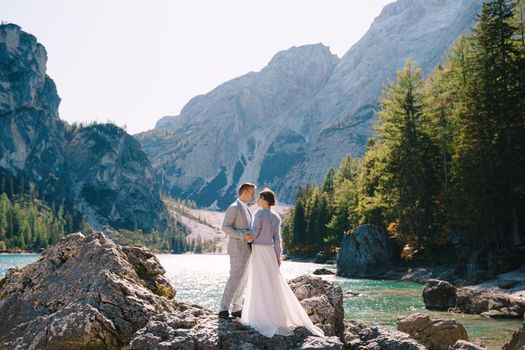 The bride and groom stand on the rocks overlooking Lago di Braies in Italy. Destination wedding in Europe, at Braies lake. In love newlyweds walk against the backdrop of amazing nature.