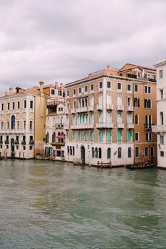 A gondola with tourists is sailing along Grand Canal amid the facades of Venetian houses standing on the water in Venice, Italy.