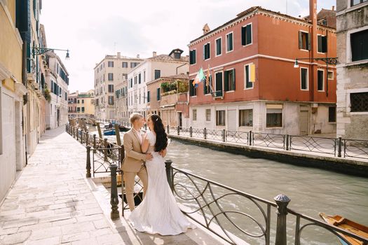 Newlyweds stand hugging on the banks of the Venice Canal. The groom hugs the bride by the waist. White wedding dress with a small beautiful train and a sandy color men's suit.