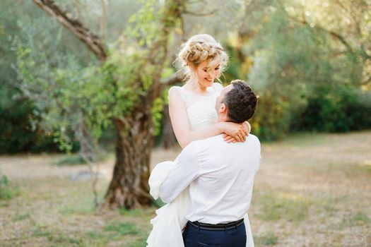 The groom circles the bride in his arms among the trees in the olive grove, the bride smiles . High quality photo