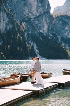 The bride and groom walk on a wooden boat dock at Lago di Braies in Italy. Wedding in Europe, at Braies lake. Newlyweds walk, kiss, cuddle against the backdrop of rocky mountains.