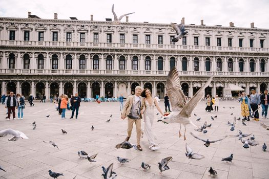 Wedding in Venice, Italy. The bride and groom are running through a flock of flying pigeons in Piazza San Marco, amid the National Archaeological Museum Venice, surrounded by a crowd of tourists.