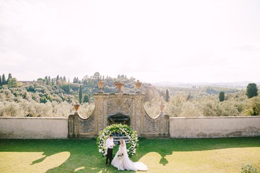 Wedding at an old winery villa in Tuscany, Italy. Wedding couple under a round arch of flowers. The groom reads out the wedding vows.
