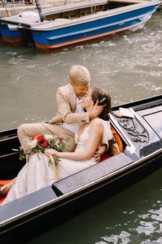 The bride and groom ride in a classic wooden gondola along a narrow Venetian canal. A close-up of cuddling newlyweds.