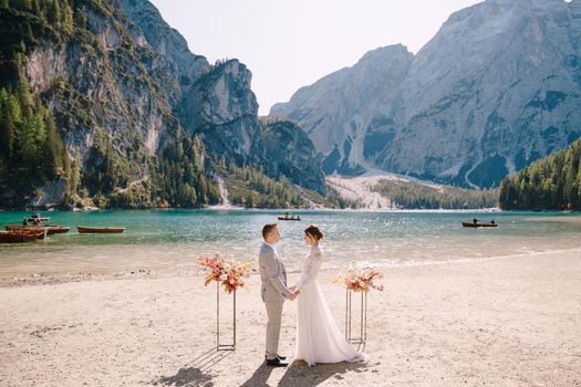 The groom dons a bride's ring, in place for the ceremony, with an arch of autumn floral columns, against the backdrop of Lago di Braies in Italy. Destination wedding in Europe, at Braies lake.