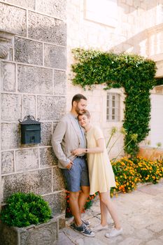 A man and a woman are embracing at the wall of a beautiful house with a window, a liana, flowers and a mailbox . High quality photo