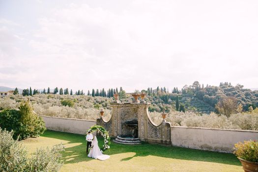 Wedding at an old winery villa in Tuscany, Italy. Wedding couple under a round arch of flowers. The groom reads out the wedding vows.