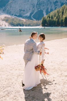 Newlyweds kiss on the spot for the ceremony, with an arch of autumn flower columns, against the backdrop of Lago di Braies in Italy. Destination wedding in Europe, at Braies lake.