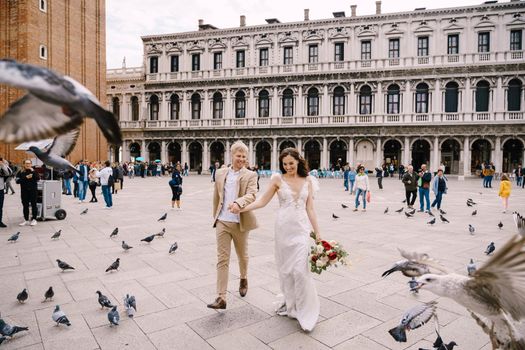 Wedding in Venice, Italy. The bride and groom are running through a flock of flying pigeons in Piazza San Marco, amid the National Archaeological Museum Venice, surrounded by a crowd of tourists.