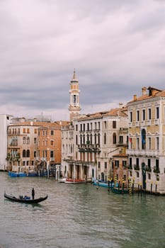 A gondola with tourists is sailing along Grand Canal amid the facades of Venetian houses standing on the water in Venice, Italy.
