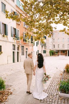 The bride and groom walk through the deserted streets of the city. Newlyweds go holding hands under a tree with yellow autumn foliage, against the facades of colorful buildings.
