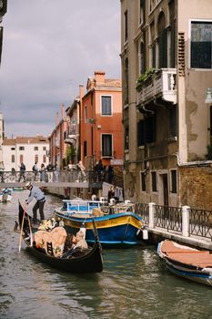 The gondolier rides the bride and groom in a classic wooden gondola along a narrow Venetian canal. Newlyweds sit in a boat and kiss.