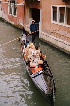 The gondolier rides the bride and groom in a classic wooden gondola along a narrow Venetian canal. Newlyweds sit in a boat and want to kiss.