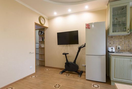 A fragment of the interior of the living room combined with the kitchen, a TV hangs on the wall, an exercise bike and a refrigerator are located nearby