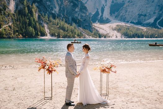 The groom dons a bride's ring, in place for the ceremony, with an arch of autumn floral columns, against the backdrop of Lago di Braies in Italy. Destination wedding in Europe, at Braies lake.