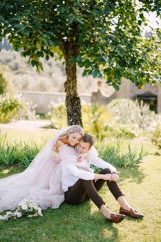 The wedding couple sits on the grass in the garden under a tree, the bride hugs the groom. Wedding in Florence, Italy, in an old villa-winery