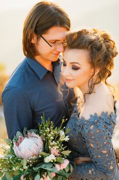 The bride and groom tenderly hug, the bride holds a bouquet with protea, eucalyptus and roses in her hands, close-up. High quality photo