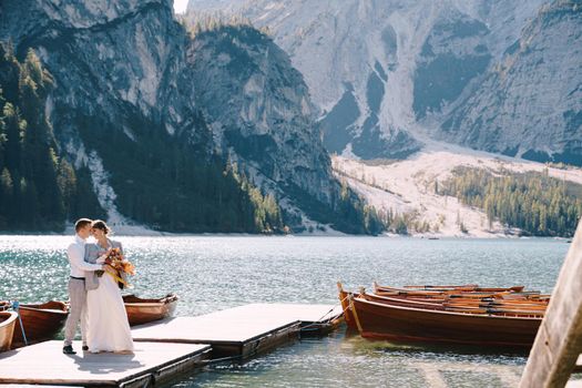 The bride and groom walk on a wooden boat dock at Lago di Braies in Italy. Wedding in Europe, at Braies lake. Newlyweds walk, kiss, cuddle against the backdrop of rocky mountains.